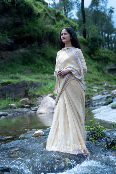 Beautiful Indian young female model wearing traditional saree standing in front of a river landscape with overcast clouds. - Foto, Bild