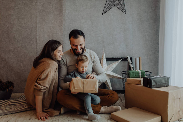 Man and woman and their child celebrate Christmas holidays together, embracing and playing with gift boxes in bright room decorated for xmas. - Photo, image