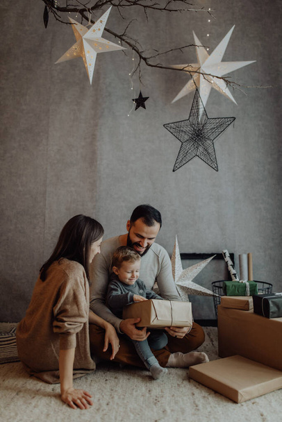 Muž a žena a jejich dítě slaví vánoční svátky společně, objímají a hrají si s dárkovými krabicemi v světlé místnosti zdobené na Vánoce. - Fotografie, Obrázek