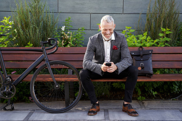 Je me repose. Homme d'affaires d'âge moyen avec succès en costume élégant textos, en utilisant le téléphone portable tout en étant assis sur le banc à l'extérieur avec un vélo à côté de lui - Photo, image