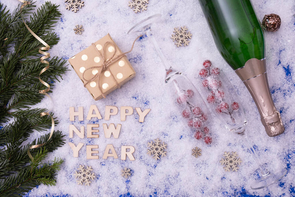 a bottle of champagne, sweets, spruce branches, a gift and wine glasses lie on the festive table next to the inscription: "Happy New Year" on a snowy background - Photo, image