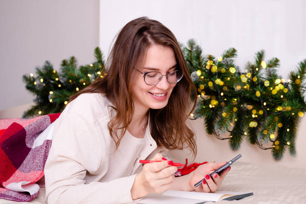 beautiful young girl makes online purchases by phone holding a credit card against the background of a christmas tree with bokeh, a shopping theme in christmas and new year - Foto, Imagem