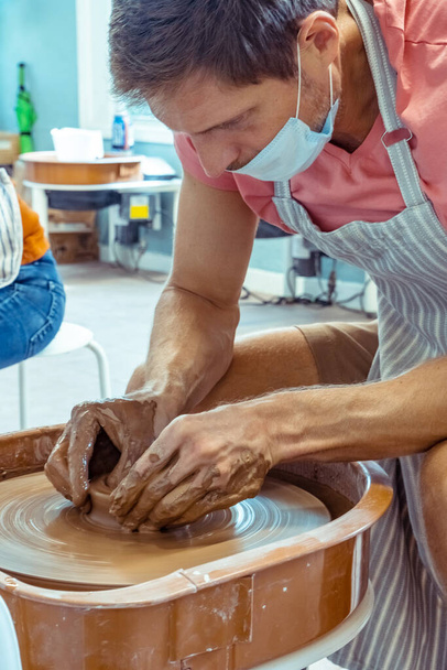 El hombre con máscara crea una taza de barro en un taller de cerámica. Pasatiempos y ocio durante la cuarentena y pandemia. - Foto, imagen