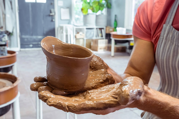 Gros plan de mains masculines sales montrant un produit fini en argile brune créé sur une roue de potier dans un atelier de poterie. Concept Hobby, leçon créative et produits faits à la main - Photo, image