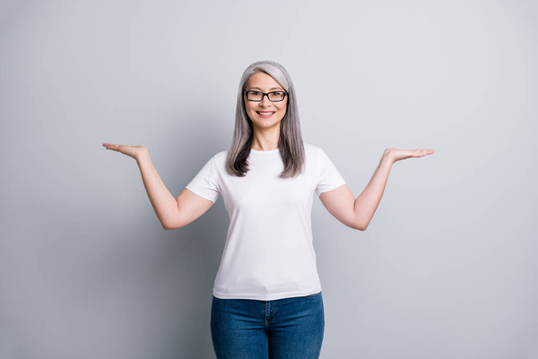 Photo of pensioner lady hold two arms empty space wear eyeglasses white t-shirt jeans isolated grey color background - Foto, Imagen