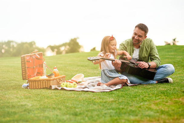 Полнометражный снимок счастливой маленькой девочки, сидящей со своим любящим отцом на зеленой траве в парке и учащейся играть на гитаре - Фото, изображение