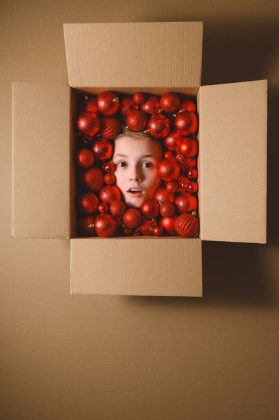 Kreative Weihnachtskomposition. Überraschtes Gesicht eines Teenagers in einem Karton mit roten Weihnachtskugeln. Ansicht von oben, vertikale Ausrichtung, Kopierraum - Foto, Bild