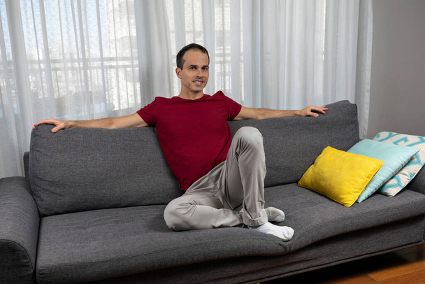Kypsä mies (44-vuotias) istuu sohvalla, avosylin värikkäiden tyynyjen vieressä. - Valokuva, kuva