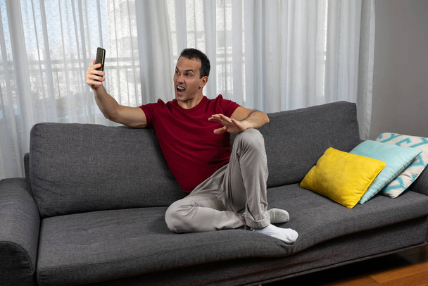 Homme mûr (44 ans) assis sur le canapé et prenant une photo de sa grimace. - Photo, image