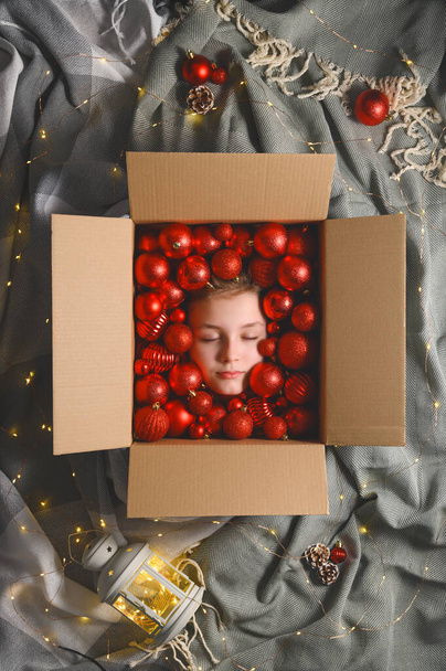 Yaratıcı Noel kompozisyonu. Ekose ve ışıkların arka planında kırmızı Noel topları olan karton kutuda uyuyan genç kızın kafası. Üst görünüm, dikey yönelim - Fotoğraf, Görsel