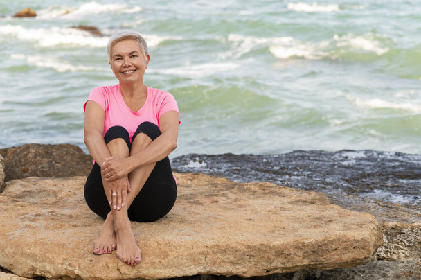 Αθλητική ώριμη γυναίκα κάθεται στην παραλία και χαμογελά - Φωτογραφία, εικόνα