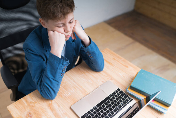 Egy kép egy kék iskolai egyenruhás kaukázusi tinédzser fiúról, aki a tanárát hallgatja laptopon távoktatás közben. - Fotó, kép
