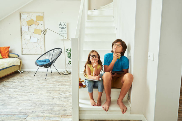 Full length shot of little brother and sister wearing eyeglasses sitting on stairs at home and thinking about something while doing homework together - Foto, immagini
