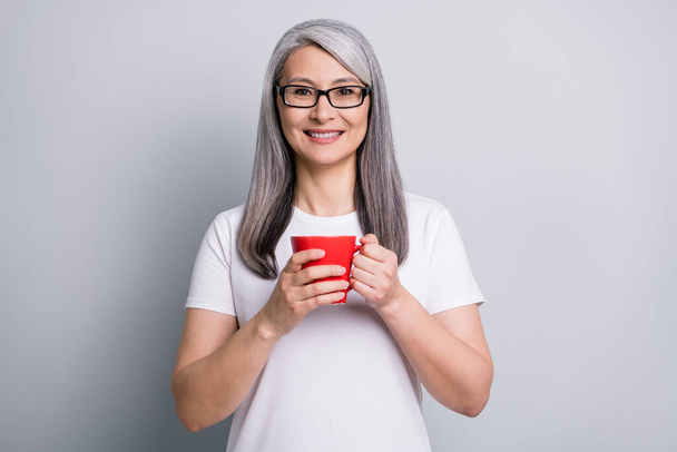 Photo of cute sweet senior lady wear casual outfit eyeglasses holding cup of tea hands arms isolated grey color background - 写真・画像