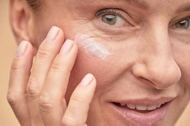 Feliz hermosa dama aplicando crema antienvejecimiento bajo los ojos - Foto, Imagen