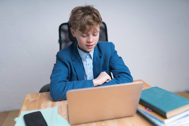 Cute Caucasian boy in blue school uniform answering questions during his online classes. He's sitting in front of his silver laptop - Foto, immagini