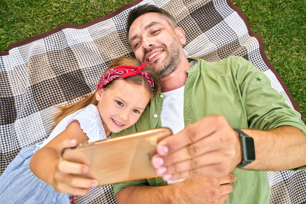 Симпатична маленька дівчинка проводить час зі своїм батьком, приймаючи селфі за допомогою смартфона, лежачи на ковдрі в прекрасному зеленому парку в теплий літній день
 - Фото, зображення