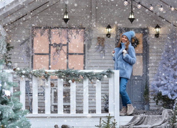 A happy girl model in a deep hat, fur coat, gloves is standing on the porch of the Christmas house! Happy New Year and Merry Christmas! - Фото, изображение