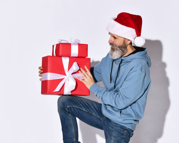 Stylový starší muž v Santově klobouku a modré mikině se dívá na vánoční červené dárky v jeho rukou. - Fotografie, Obrázek