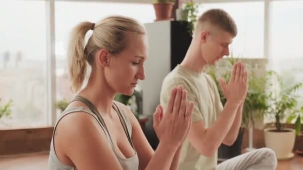 Μέτριο κοντινό πλάνο των νέων χαλαρό ζευγάρι που έχουν πρακτική γιόγκα κάνει στάση προσευχή κάθεται με τα μάτια κλειστά και τα πόδια σταυρωμένα - Πλάνα, βίντεο
