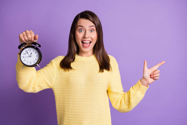 Photo portrait of positive woman holding clock with one hand pointing finger to side with open mouth isolated on vivid purple colored background - Fotó, kép