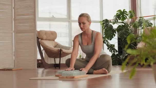 Full shot of woman sitting with legs crossed placing palm on sadhu board with sharp metal nails at modern yoga practice - Footage, Video