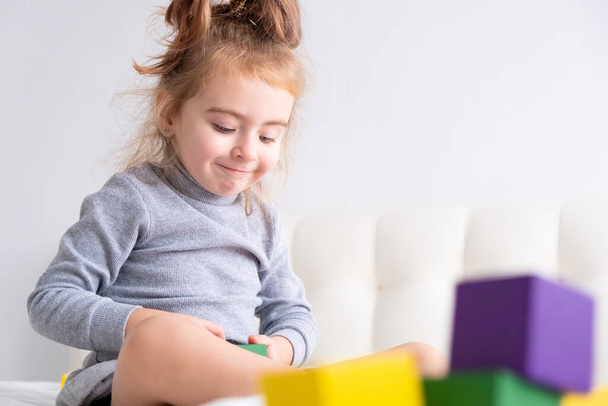 Маленька дівчинка грає з різнокольоровими дерев'яними кубиками, сидячи на ліжку. Домашні заходи для дітей
. - Фото, зображення
