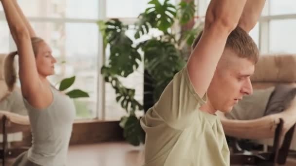 Medio primo piano del giovane uomo caucasico con il suo partner femminile che fa posa piccione addormentato yoga piegandosi in avanti dalla posizione seduta in studio moderno con finestre panoramiche - Filmati, video