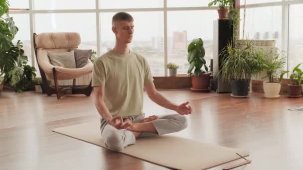 Full shot de joven concentrado en pose de loto haciendo mudra especial de postura de dedo, tomando respiración profunda preparándose para la meditación de yoga en interiores - Imágenes, Vídeo
