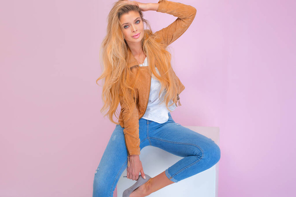 Beautiful sexy blonde girl with long hair and tanned body wearing brown jacket and blue jeans, sitting and sensual posing at pink wall. - Foto, afbeelding