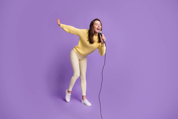 Foto de cuerpo entero de una estudiante morena sosteniendo el micrófono cantando con los ojos cerrados sonriendo aislada sobre fondo de color púrpura vivo - Foto, imagen