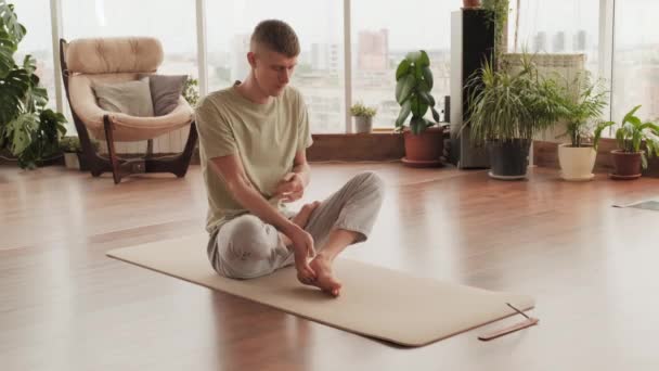 Tiro completo de jovem caucasiano magro no mapa de ioga fazendo postura de lótus completo se preparando para meditar sozinho com os olhos fechados - Filmagem, Vídeo