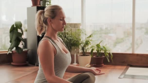 Średnie zbliżenie młodej skoncentrowanej kobiety siedzącej z nogami skrzyżowanej oddychającej głęboko i medytującej w jasnym studio jogi - Materiał filmowy, wideo