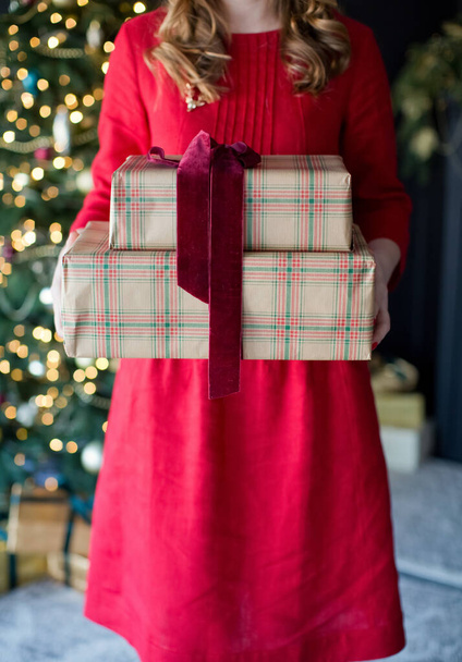 Žena v červených šatech drží vánoční dárkové krabice s stuhou u vánočního stromečku. Vánoční dárky, vánoční koncept - Fotografie, Obrázek