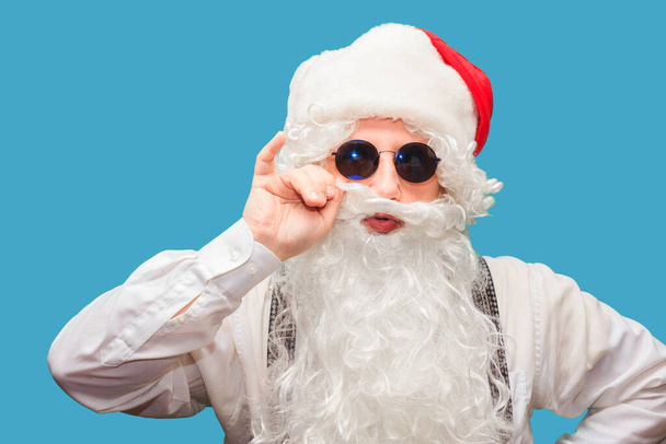 Winter fashion and sales. Portrait of a handsome man in a Santa hat, winking while standing against a red background. The hipster is holding glasses and looking at the camera. Place for text. - Foto, Imagem