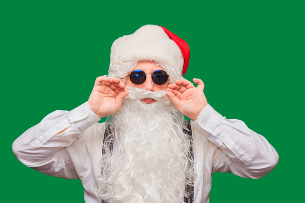 Winter fashion and sales. Portrait of a handsome man in a Santa hat, winking while standing against a red background. The hipster is holding glasses and looking at the camera. Place for text. - 写真・画像