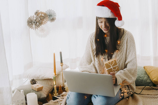 Ευτυχισμένη νεαρή γυναίκα αγοράζουν δώρα σε απευθείας σύνδεση στο laptop κάθεται στην αίθουσα Boho με χριστουγεννιάτικα δώρα και εορταστικές διακοσμήσεις. Κομψό θηλυκό σε φώτα και καπέλο santa σε βιντεοκλήση με την οικογένεια - Φωτογραφία, εικόνα