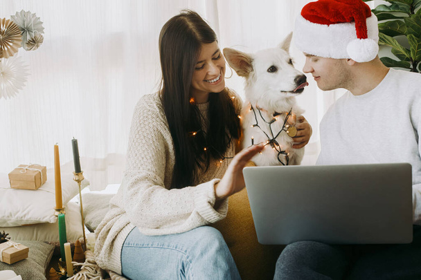 Szczęśliwa, młoda rodzina z uroczym psem machającym rękami w rozmowie wideo na laptopie w pokoju boho z prezentami świątecznymi i wystrojem. Stylowa para w świetle i Santa kapelusz świętowanie wakacji z rodziną online - Zdjęcie, obraz