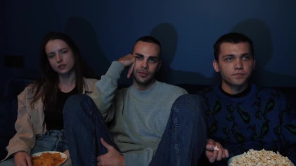 Three bored friends watching tv sitting on couch and eating popcorn, boring movie, show - Video
