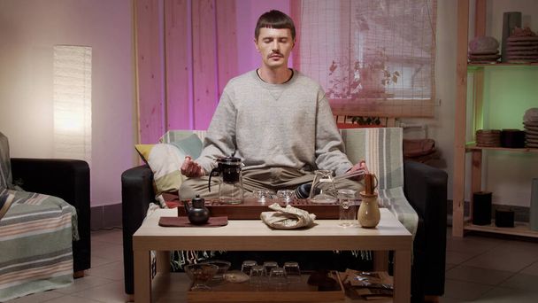 Ein junger Mann meditiert in Lotusposition auf einem Sofa während einer Teezeremonie in einem bunten Wohnzimmer - Foto, Bild