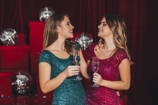 Dos niñas felices alegres que tienen una fiesta de año nuevo, celebrando la Navidad. Beben champán y abren regalos. Llevan vestidos sexy que curva su cuerpo de manera sexy. Muchos regalos rojos en la espalda. - Foto, imagen