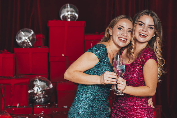 Dwie radosne, szczęśliwe dziewczyny urządzające przyjęcie noworoczne, świętujące święta. Piją szampana i otwierają prezenty. Noszą seksowne sukienki, które podkręcają ich ciało w seksowny sposób. Wiele czerwonych prezentów na plecach. - Zdjęcie, obraz