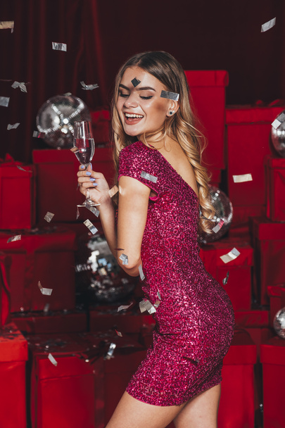 Веселая счастливая девушка, празднующая Рождество на новогодней вечеринке. Она пьет шампанское и открывает подарки. Она носит сексуальные платья, которые изгибают их тело сексуально. Много красных подарков сзади - Фото, изображение