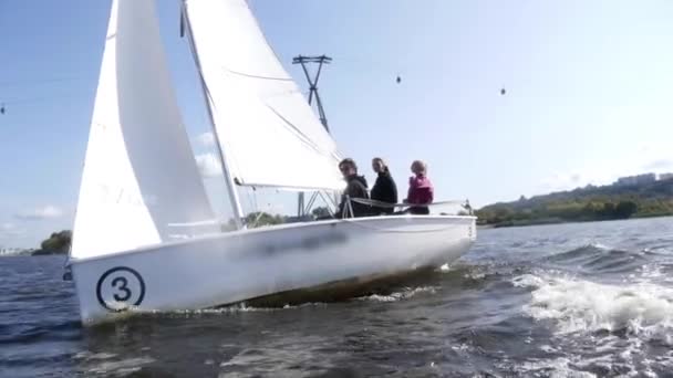 Op een wit jacht met een wit zeil drijven drie jongeren in slow motion op de rivier onder de kabelbaan. - Video