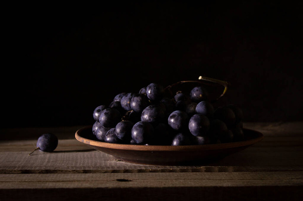 Ripe bunch of red grapes on a clay plate on a wooden rustic table, shot in low key - Foto, Bild