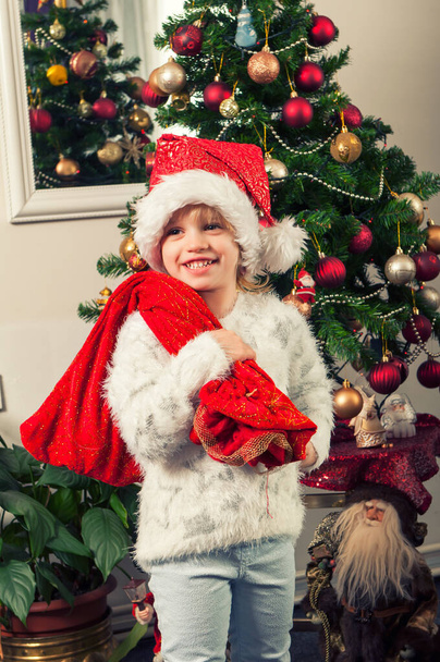 Beautiful little girl with santa claus hat and gift bag standing beside christmass tree - Foto, afbeelding