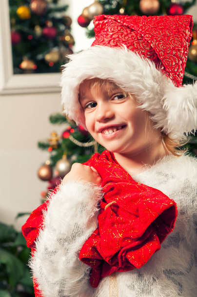 Beautiful little girl with santa claus hat and gift bag standing beside christmass tree - Foto, Imagem