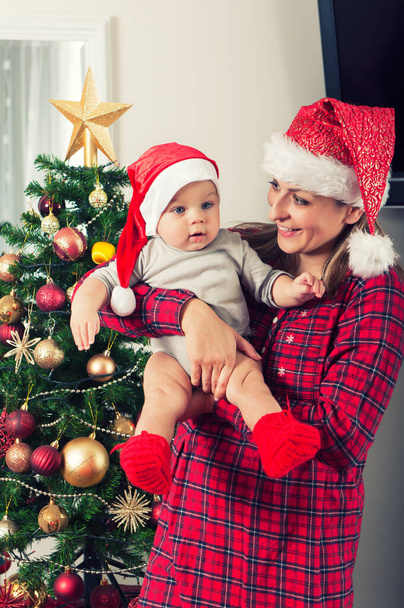 Mother and her beautiful baby boy posing beside christmass tree with santa claus hats - Foto, Imagen
