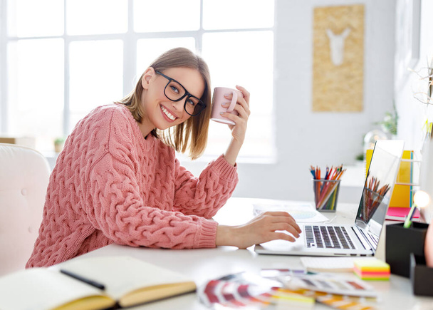 Вид сбоку на восхищенную молодую женщину, улыбающуюся и смотрящую на камеру через плечо во время использования ноутбука для работы над дизайнерским проектом на hom - Фото, изображение
