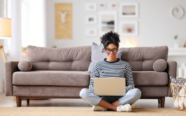 Vrolijke jonge etnische vrouwelijke freelancer in bril en casual kleding gericht op het scherm en interactie met laptop, terwijl alleen zitten op de vloer in het licht van de moderne living roo - Foto, afbeelding